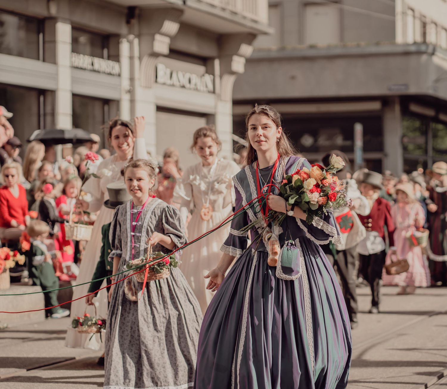 Women in elegant historical gowns carry colorful flower bouquets during a festive street parade.