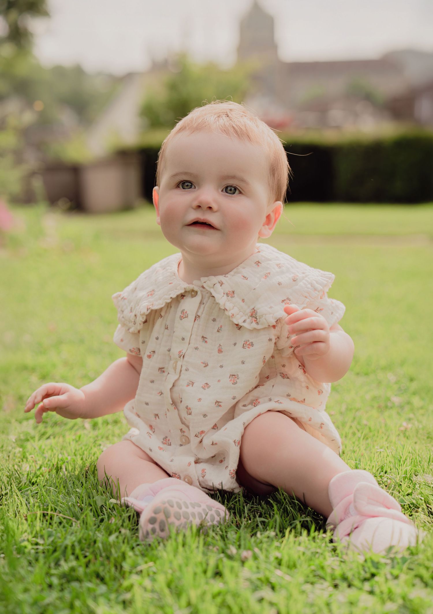 Adorable baby in cream floral ruffle romper with pink socks sitting on green grass outdoors.