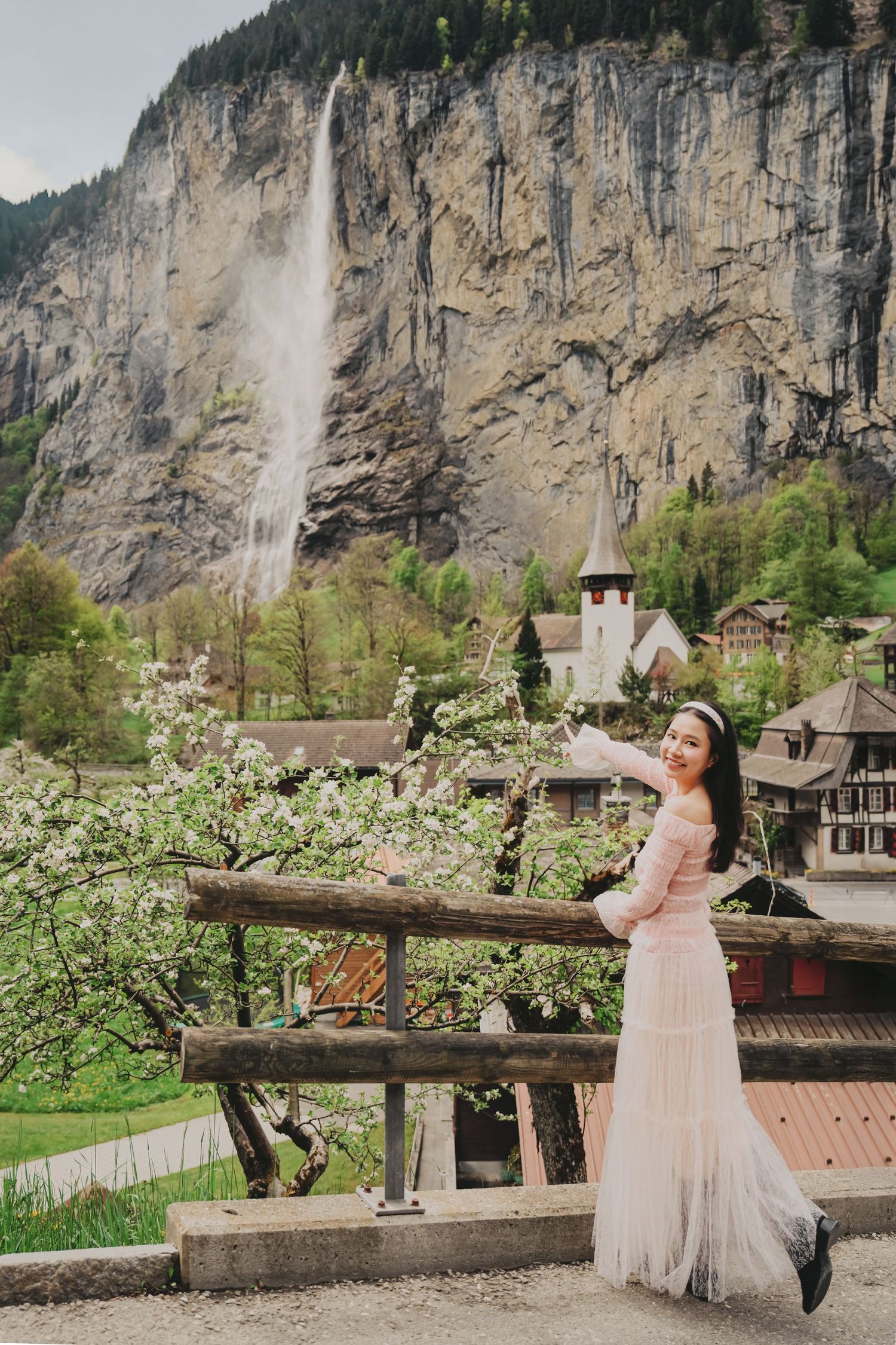 Woman in pink dress posing by wooden fence with Staubbach waterfall and Lauterbrunnen village in background.