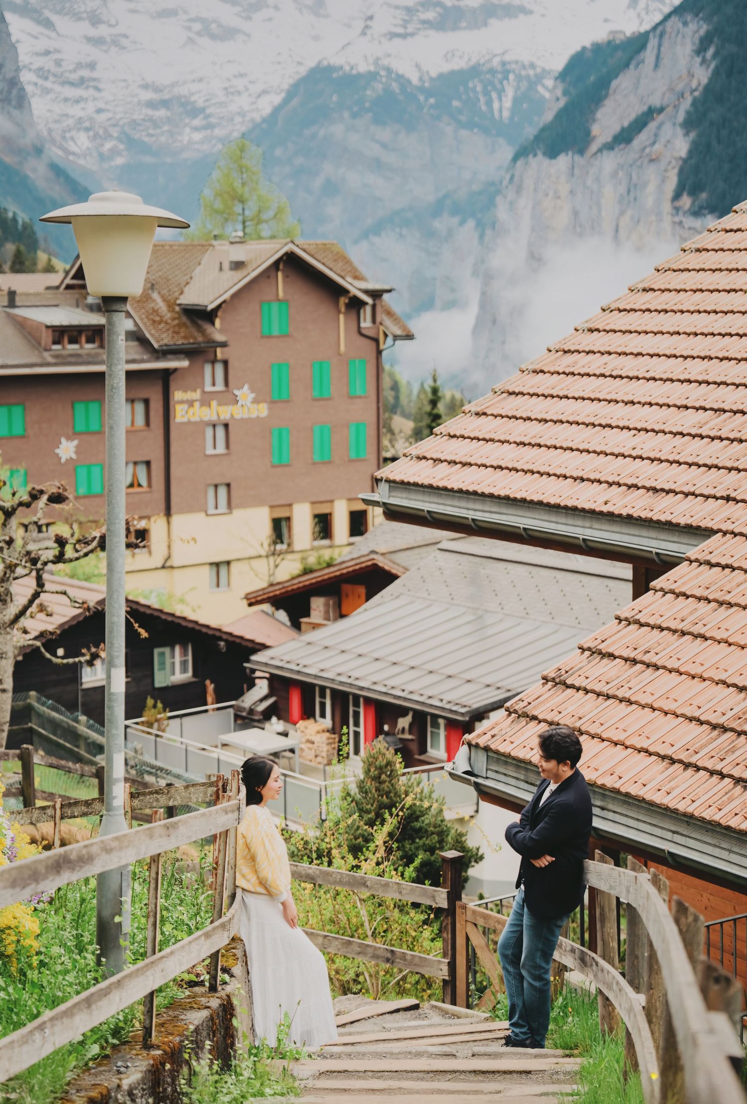Couple on wooden staircase with scenic Swiss Alpine village and waterfall backdrop.