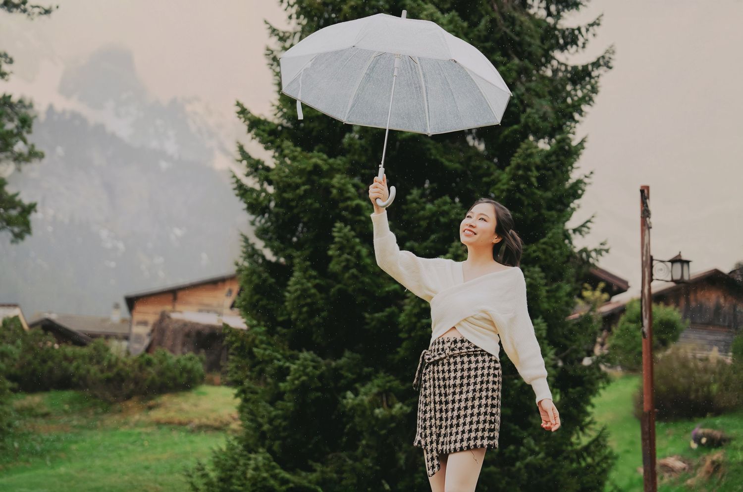 Smiling woman lifting white umbrella joyfully in green alpine meadow with mountain backdrop.