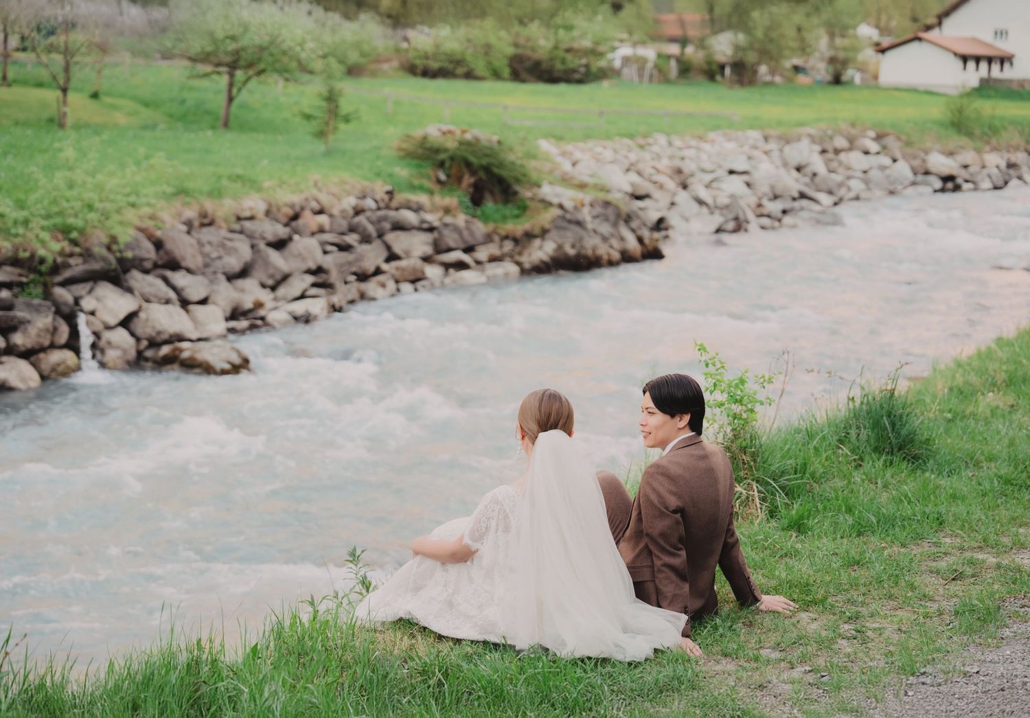 Bride and groom sit together on grassy riverbank gazing at each other in a romantic alpine setting.