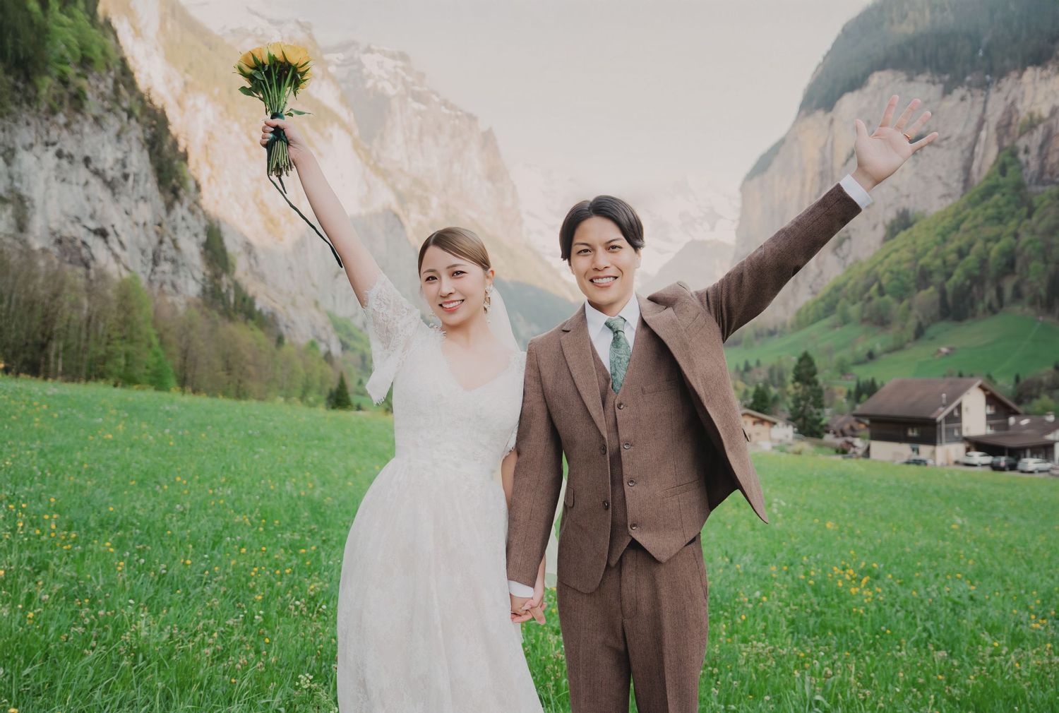 Celebrating newlyweds raising arms with yellow bouquet in scenic Swiss Alpine meadow.