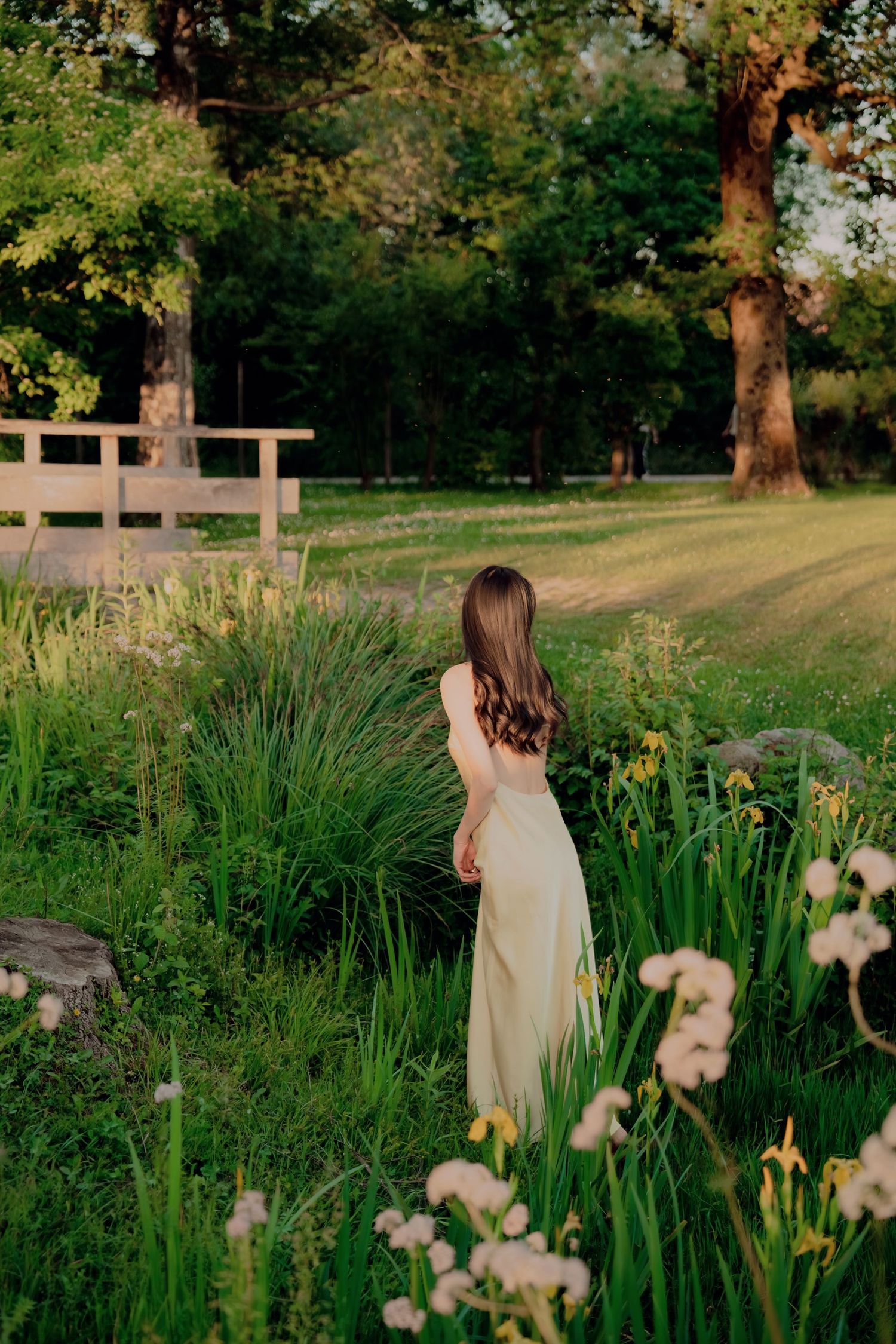 Young woman in flowing cream dress standing among wildflowers and iris blooms in lush green garden at golden hour.