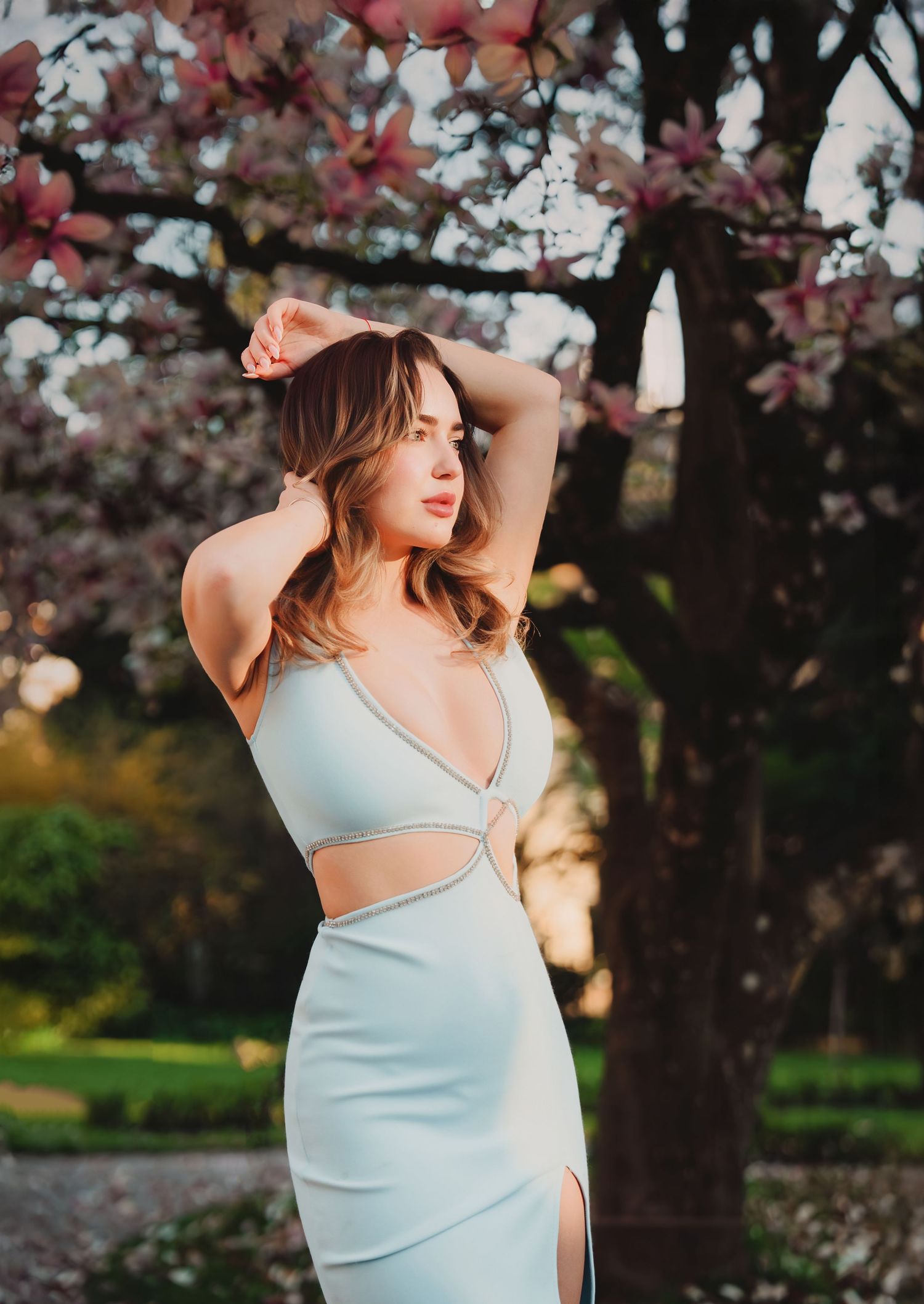Woman in light blue cutout dress posing near blooming magnolia tree in soft evening light.