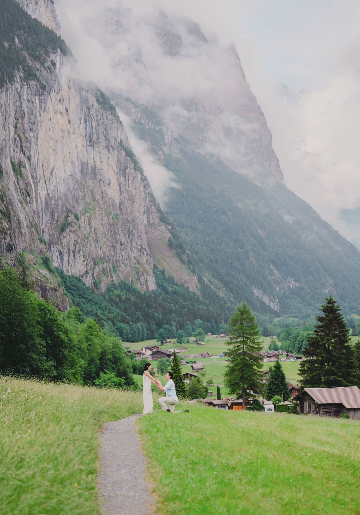 Couple holding hands on a mountain path with a stunning Swiss alpine village and misty cliffs.