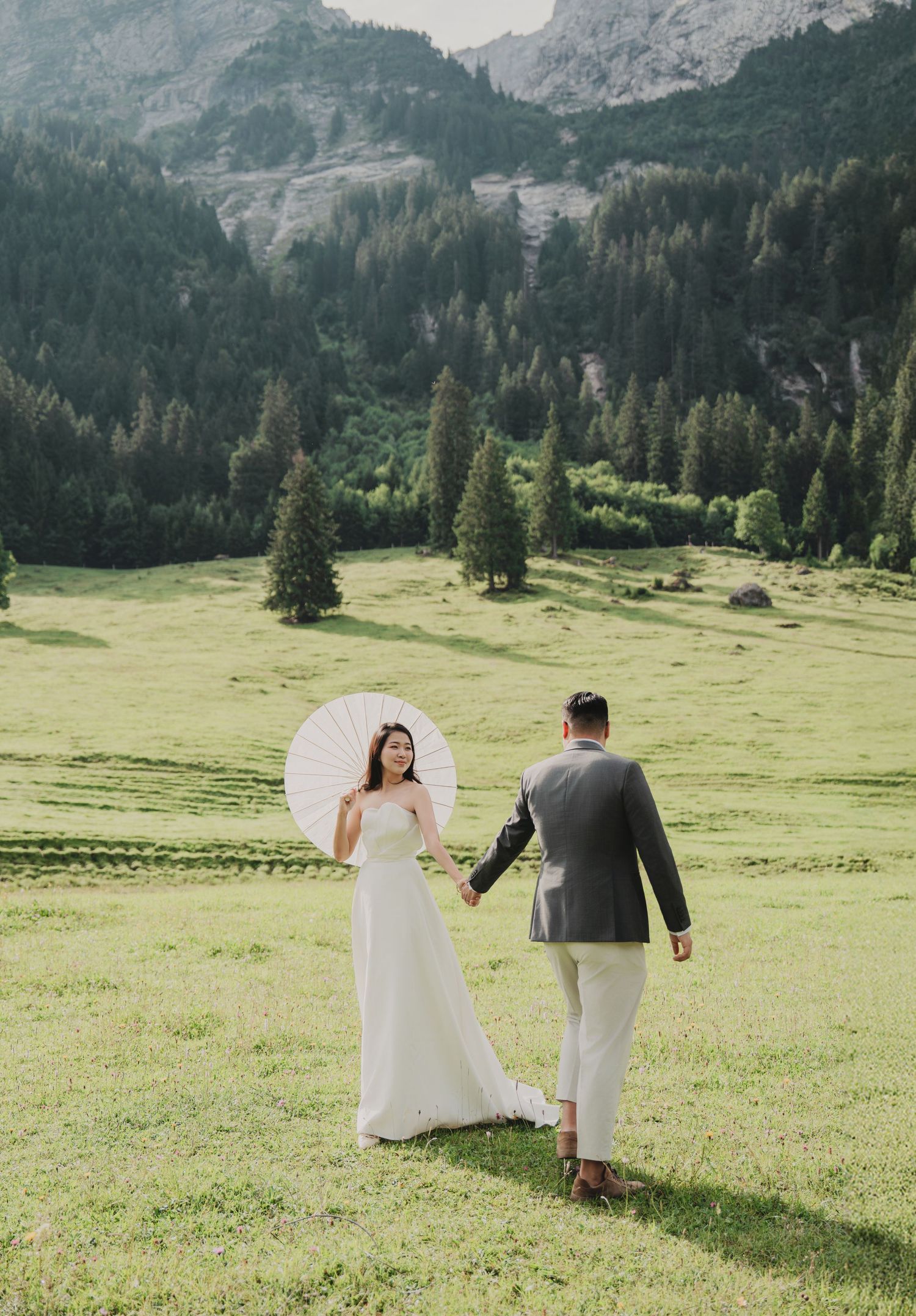 Bride in white gown holding parasol walks hand in hand with groom through alpine meadow with mountain backdrop.