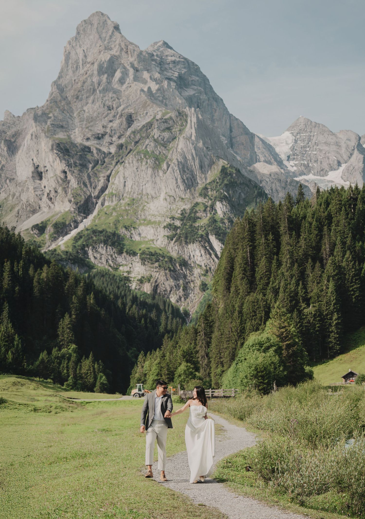 Bride in white gown and groom in gray suit strolling along a scenic alpine valley path with majestic mountains.