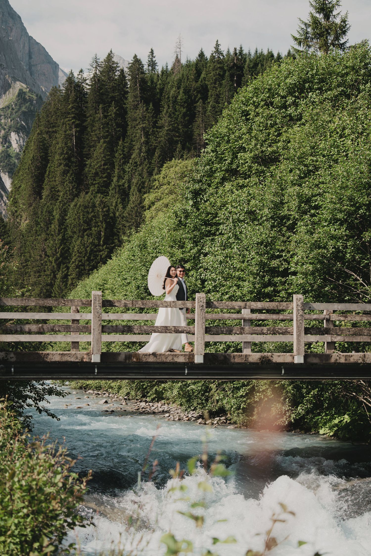 Wedding couple embracing on a wooden bridge over a turquoise Alpine river surrounded by dense forest.