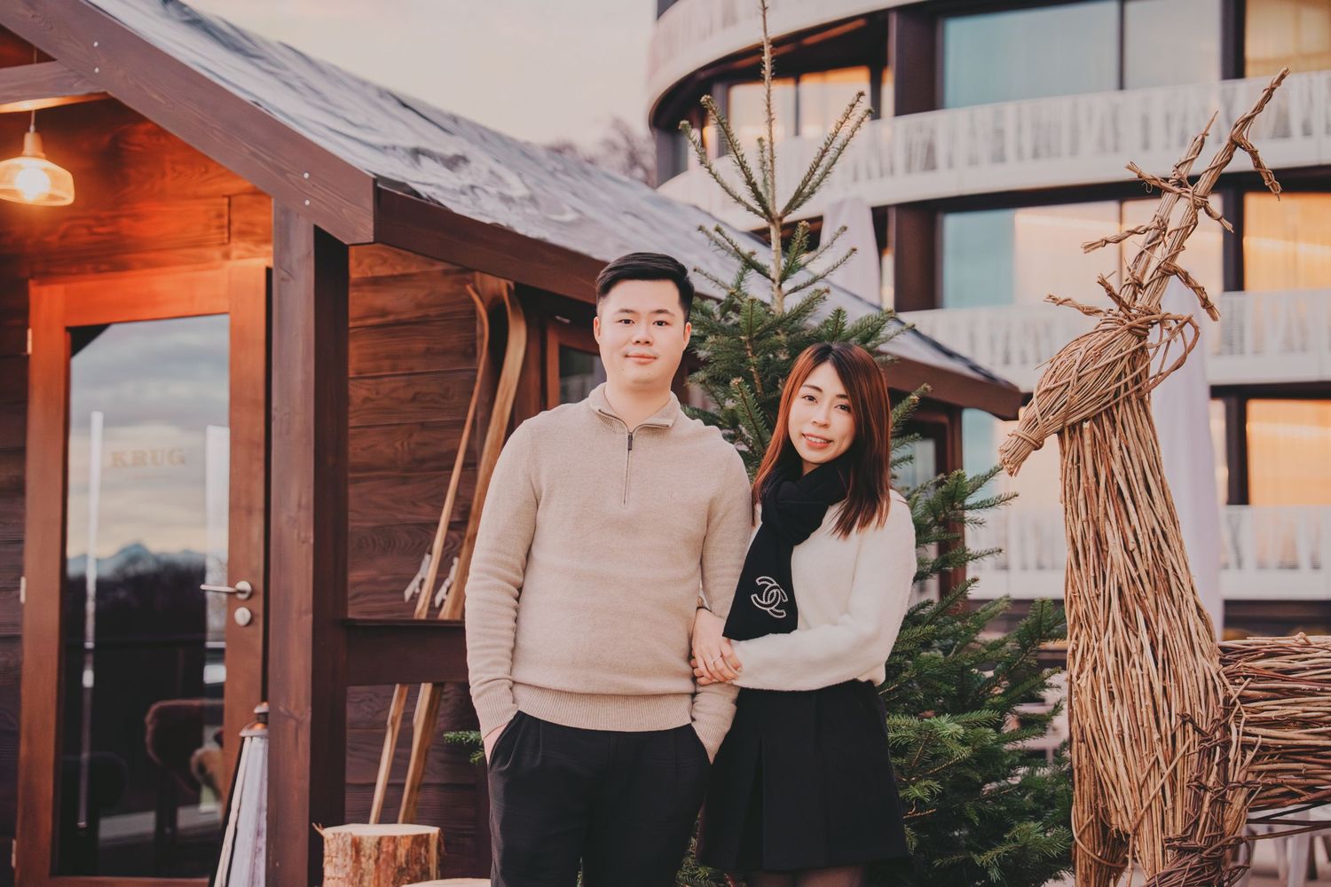 Couple posing together outside a wooden chalet with Christmas decorations at dusk.