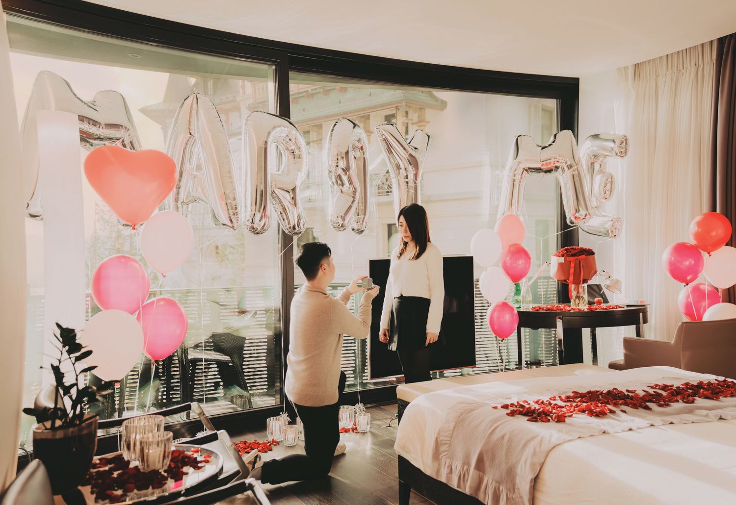 Man on one knee proposing in hotel room decorated with rose petals, balloons, and silver balloons.