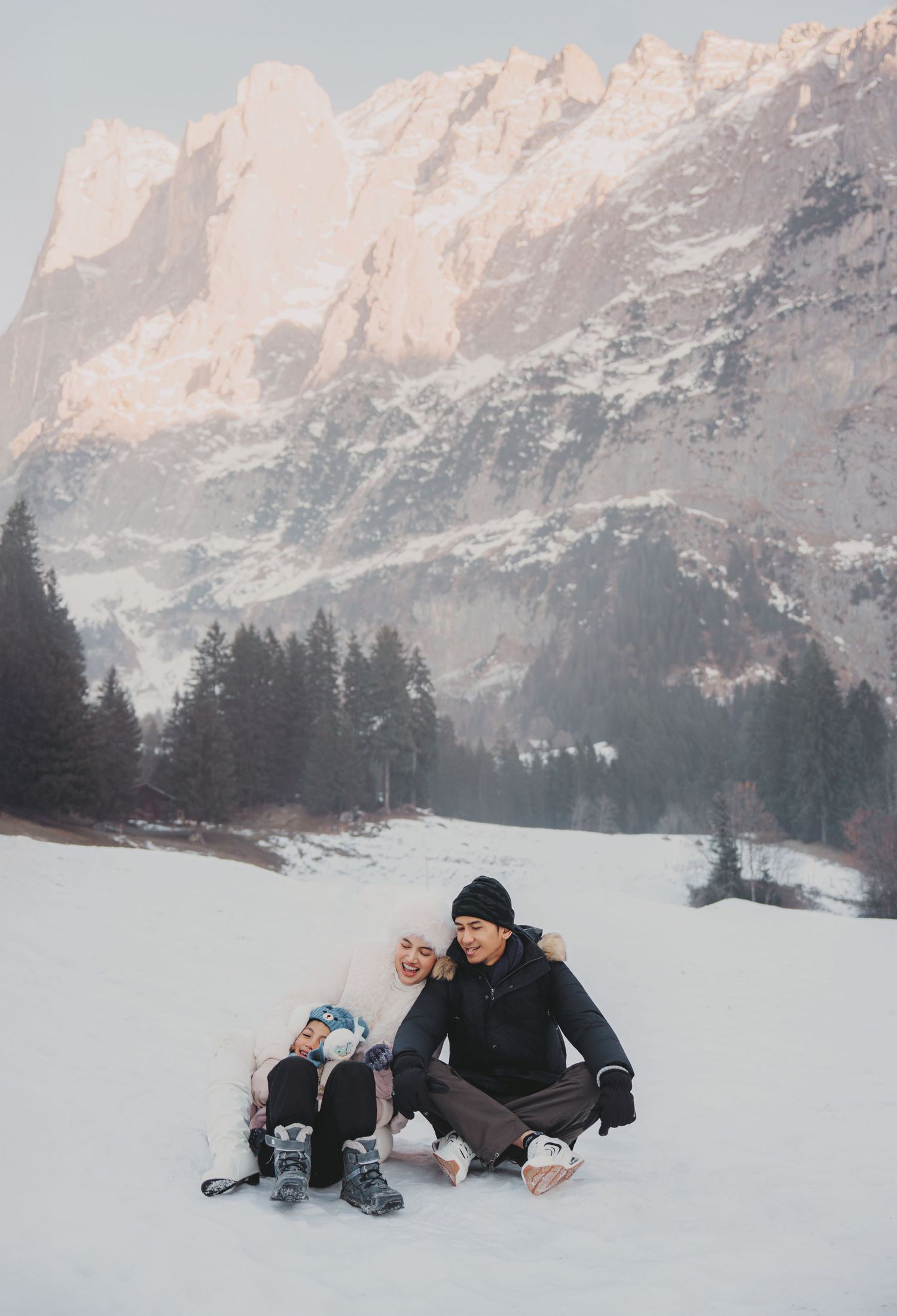 Couple sitting together on snowy alpine meadow with dramatic mountain peaks and pine forest backdrop in winter.