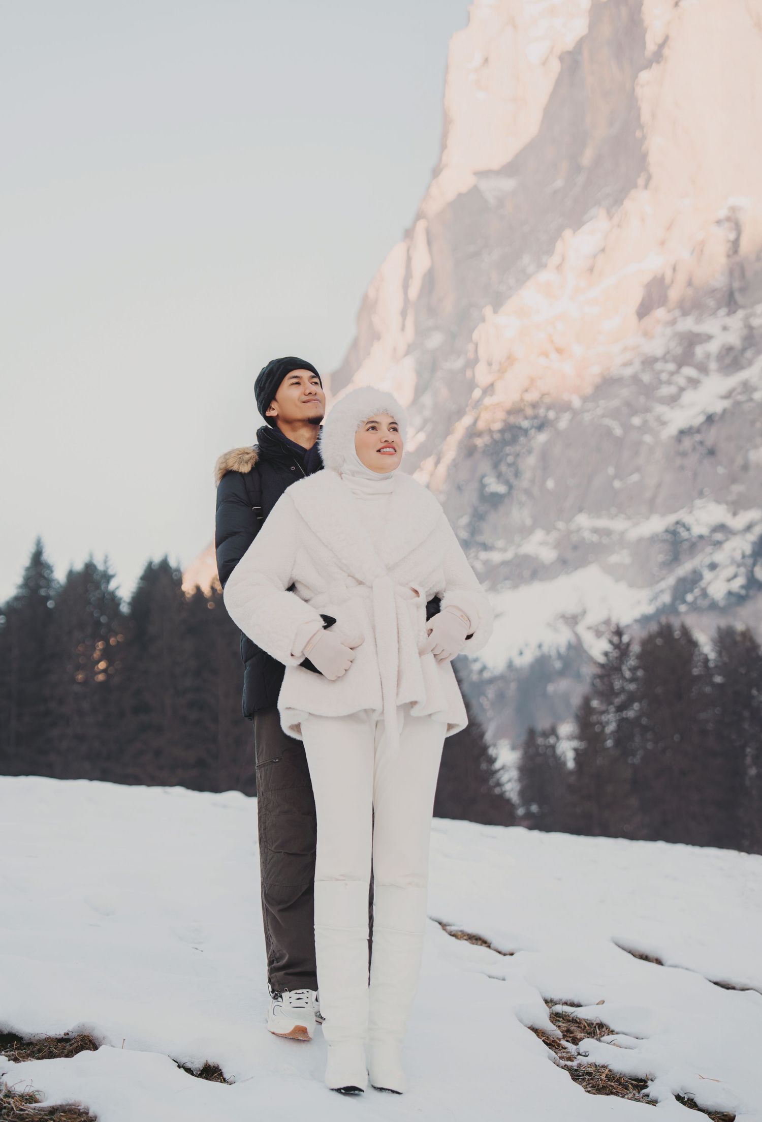 Couple in white winter outfits standing together gazing at sunlit rocky Alpine mountain in snow.