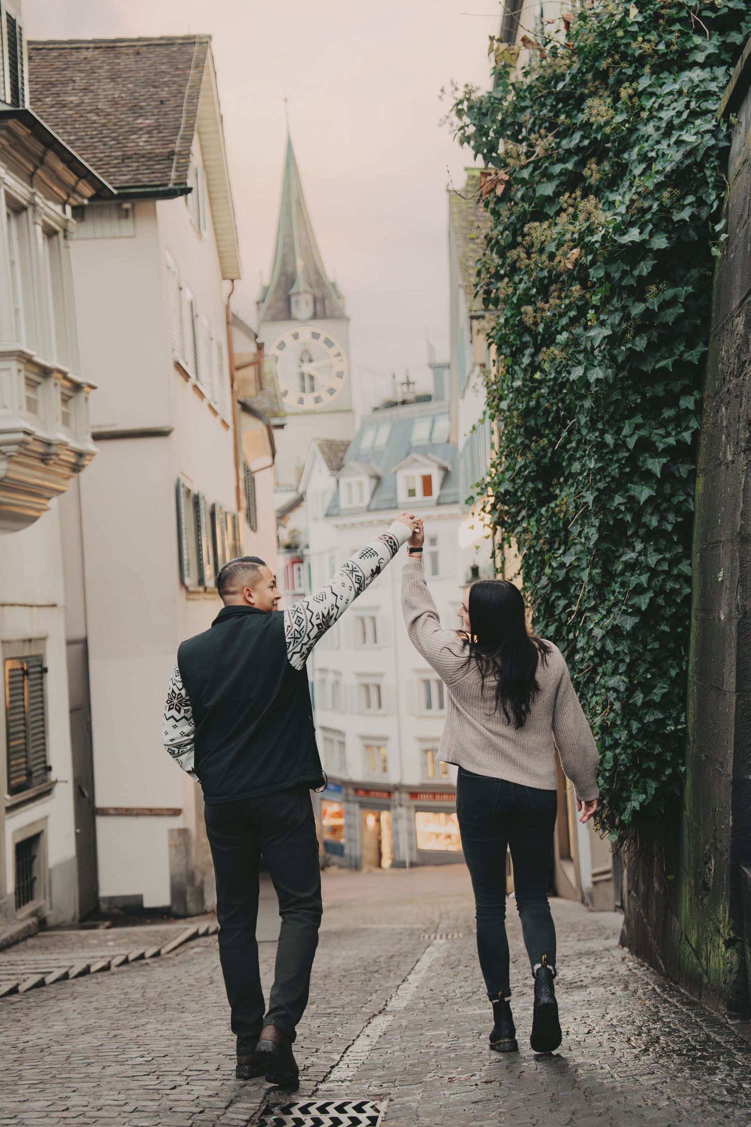 Couple raising hands joyfully while walking through a scenic European old town street.
