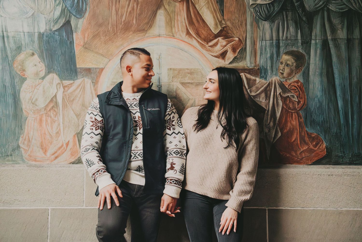 Couple holding hands and smiling at each other in front of a classical fresco mural.