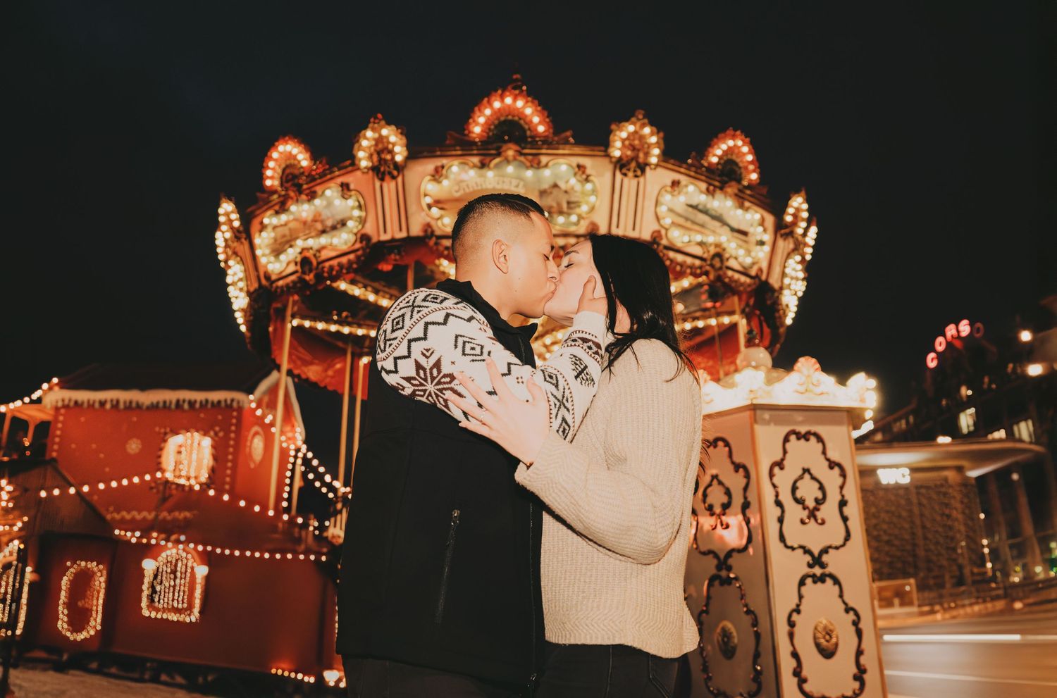 Couple sharing a kiss in front of a glowing vintage carousel at a festive Christmas night market.