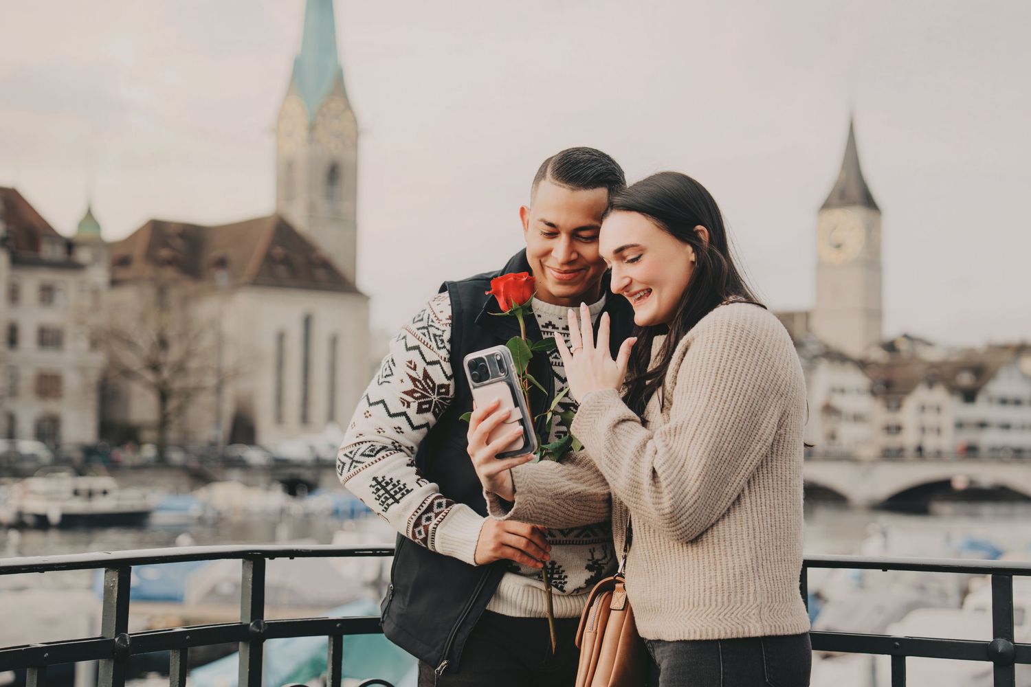 Couple smiling at phone holding red rose with Zurich skyline in background.