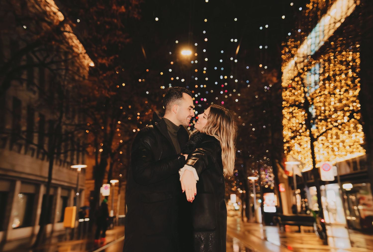 Couple sharing a romantic moment on a festive city street illuminated with golden Christmas lights at night.