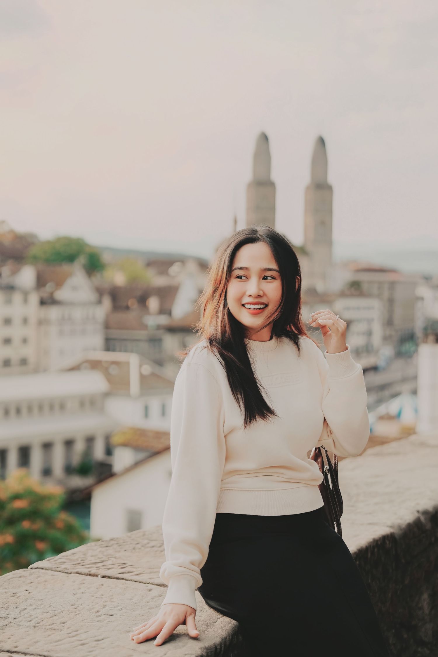 Smiling Asian woman in white sweater sitting on stone wall with Grossmünster towers behind her.