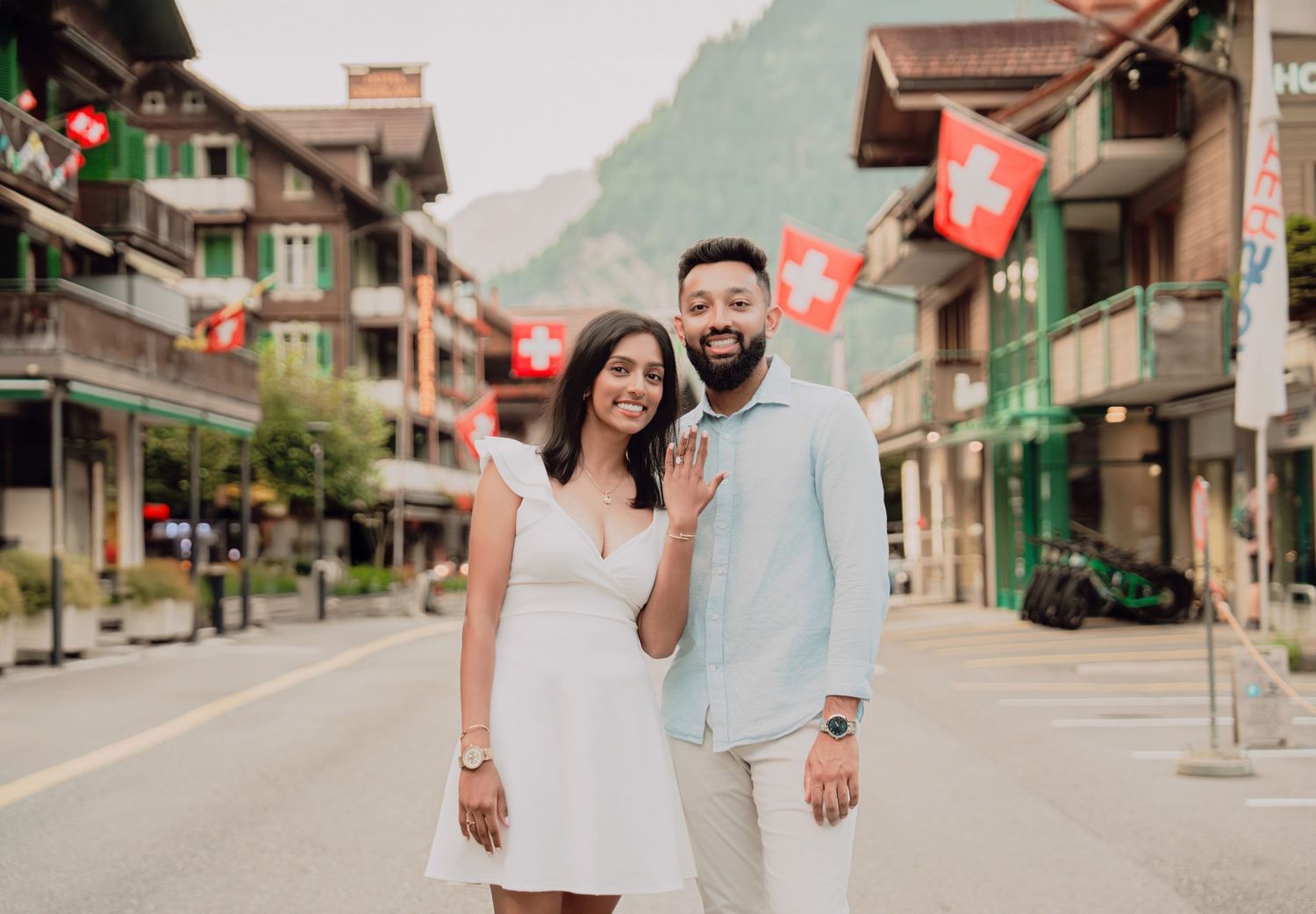 Happy couple smiling on a Swiss street in Lauterbrunnen, surrounded by traditional buildings and Swiss flags.