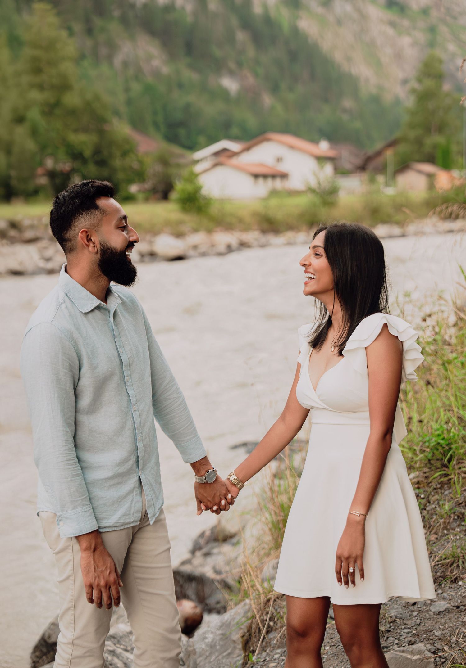 Happy couple holding hands and smiling at each other beside a river in a scenic Swiss mountain valley.
