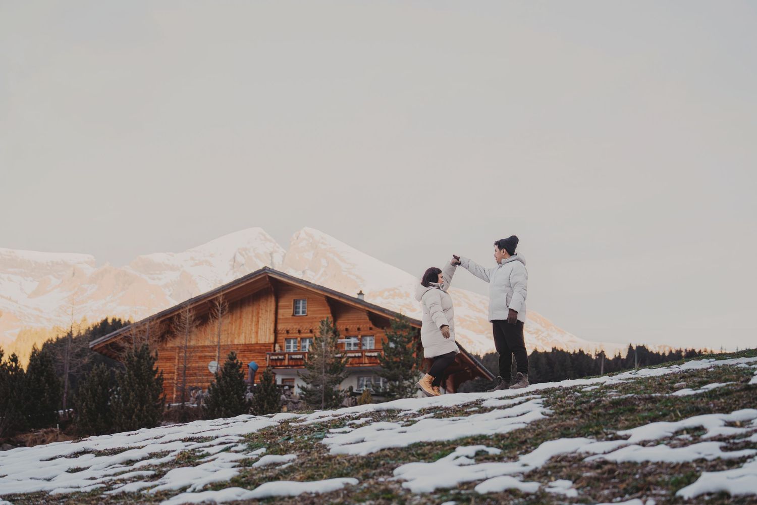 Couple dancing near rustic wooden chalet on snowy hillside at sunset with mountain backdrop.