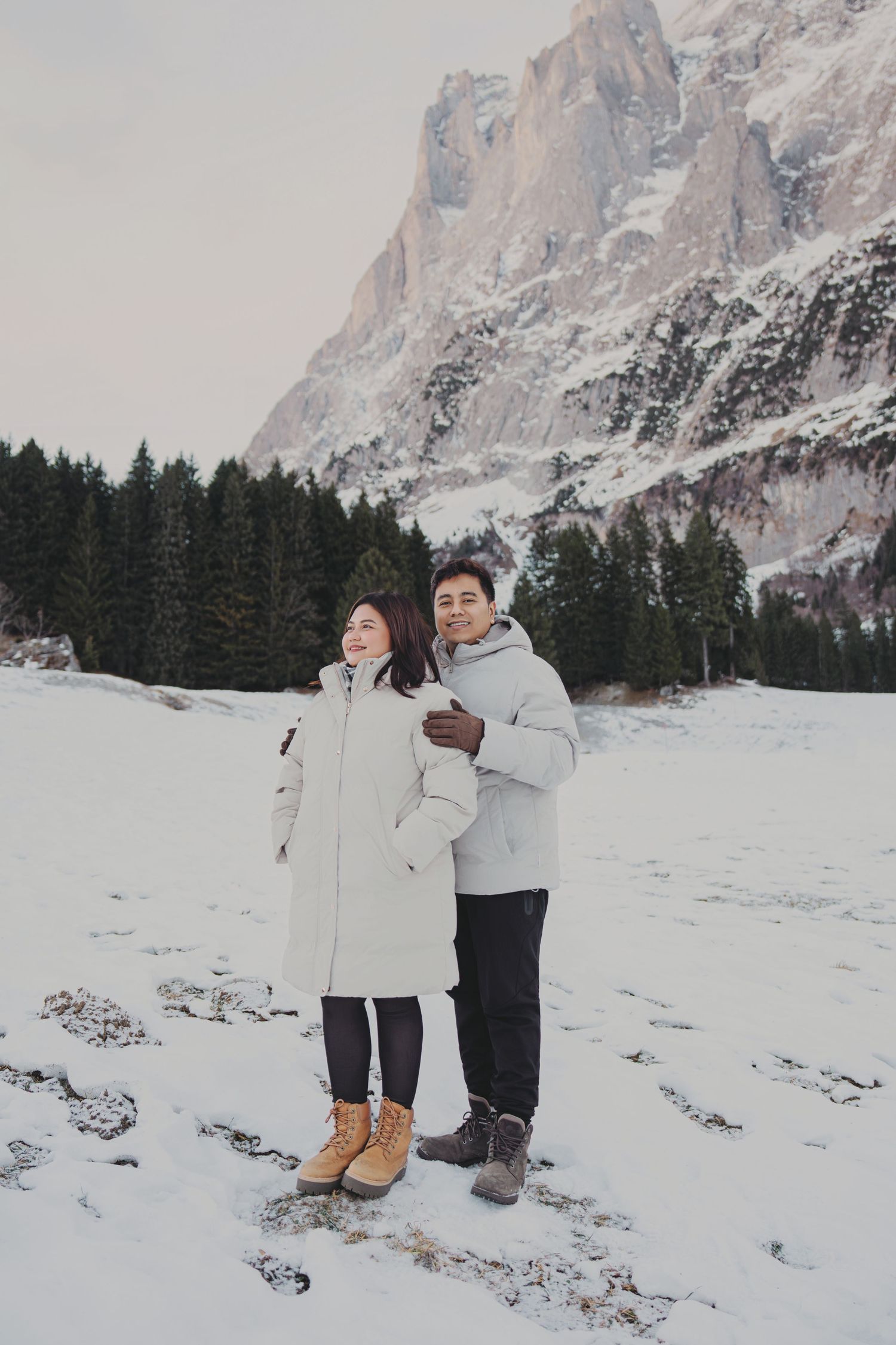 Couple in white coats embracing in snowy field with dramatic rocky mountain and pines.