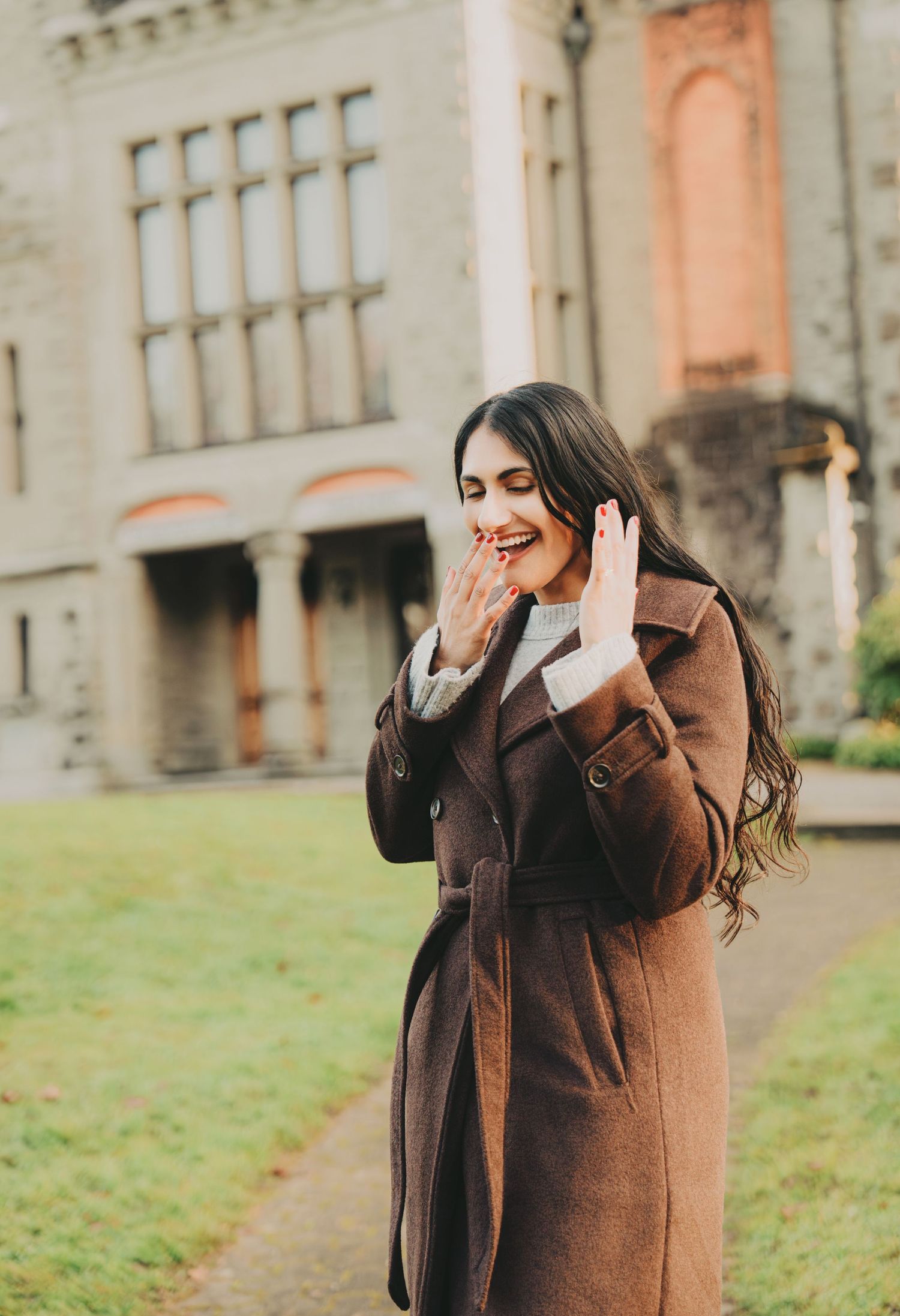Smiling woman in brown coat showing engagement ring outside a historic stone building.