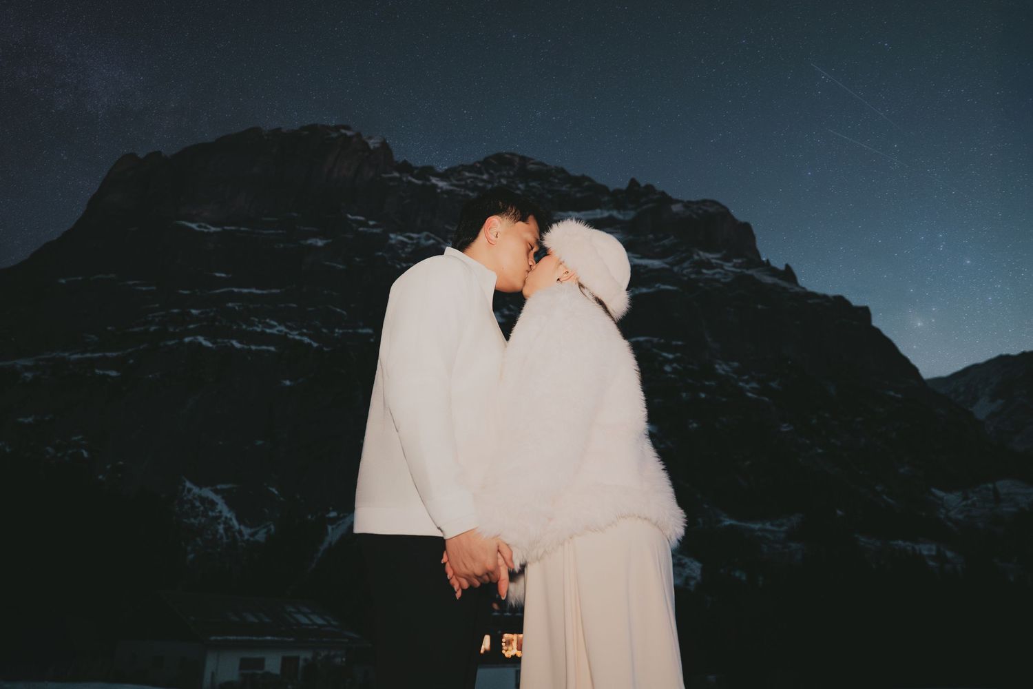 Couple sharing a kiss in matching white outfits under starry mountain night sky.
