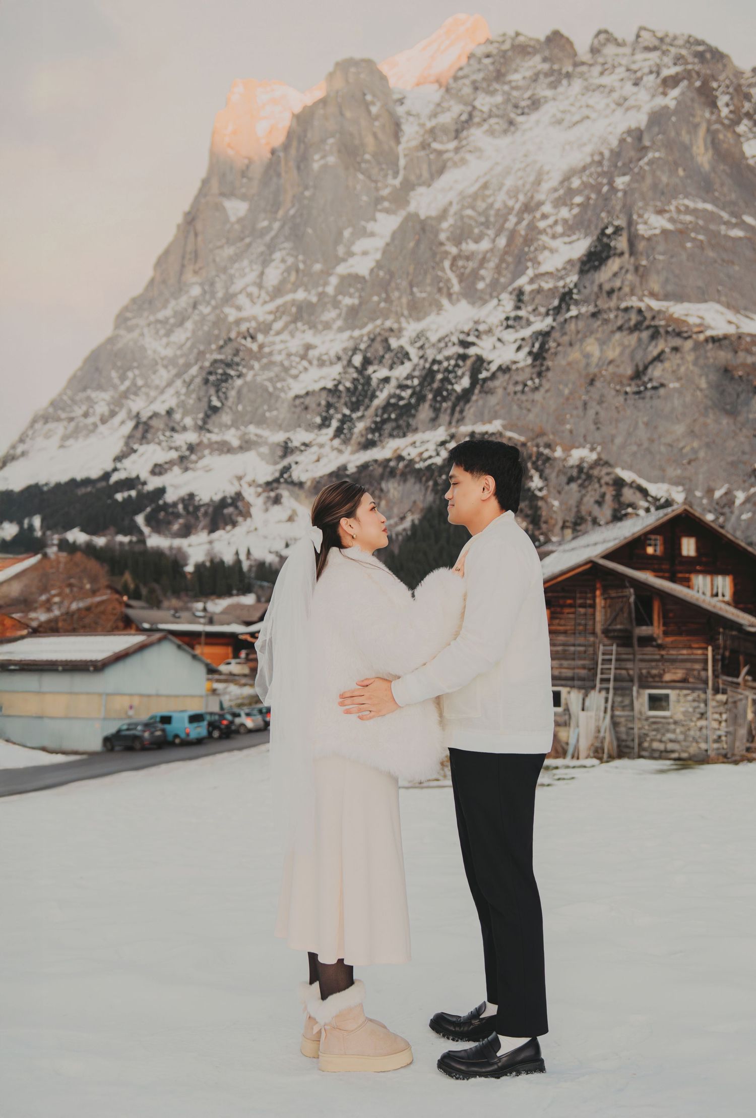 Romantic couple embracing face-to-face in white attire against snow-covered Swiss Alps at sunset.