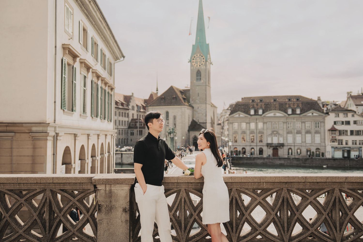 Couple sharing a moment on a bridge in Zurich old town with Fraumunster church spire in background.