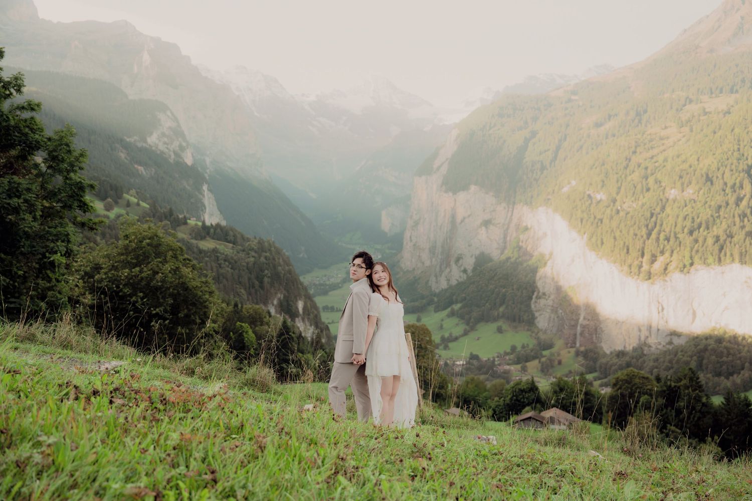Couple in wedding attire standing together on a green hillside with a dramatic misty Alpine valley behind them.