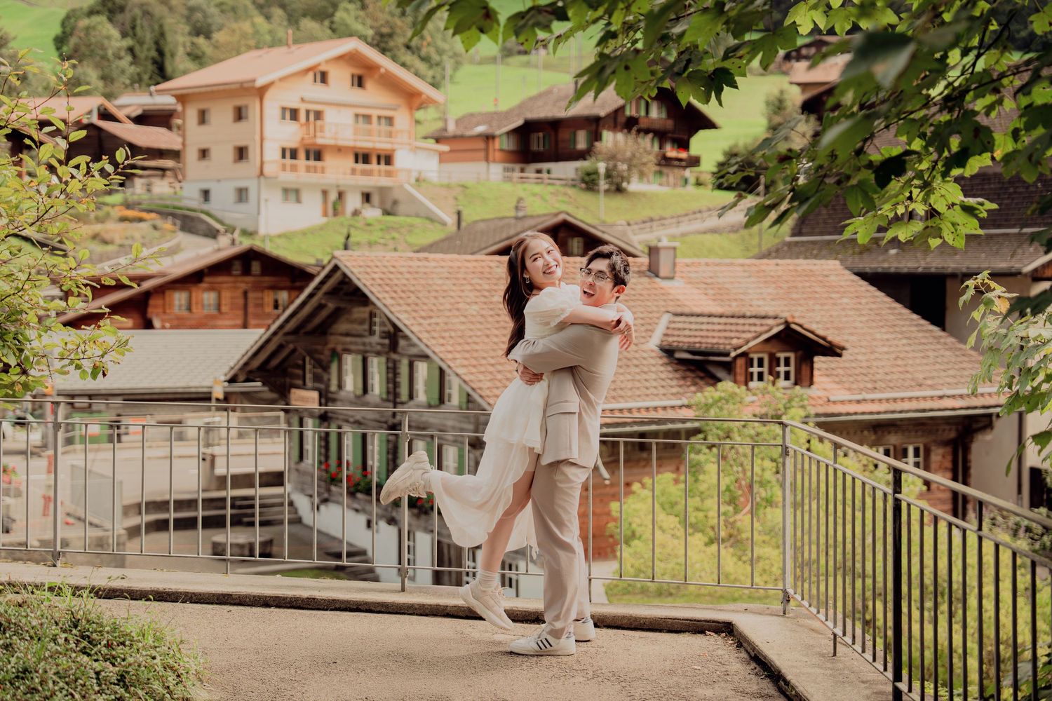 Groom lifting bride joyfully on a terrace with Swiss alpine village homes behind them.