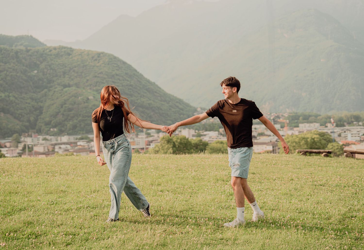 Young couple walking hand in hand across green field with mountain town behind.