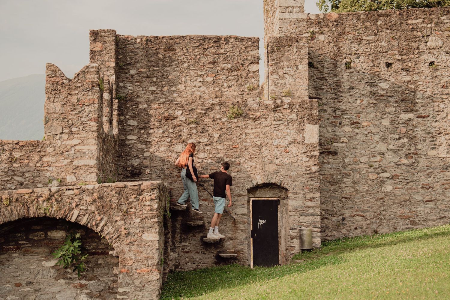 A couple climbing stone steps along the exterior wall of a historic medieval castle ruin.