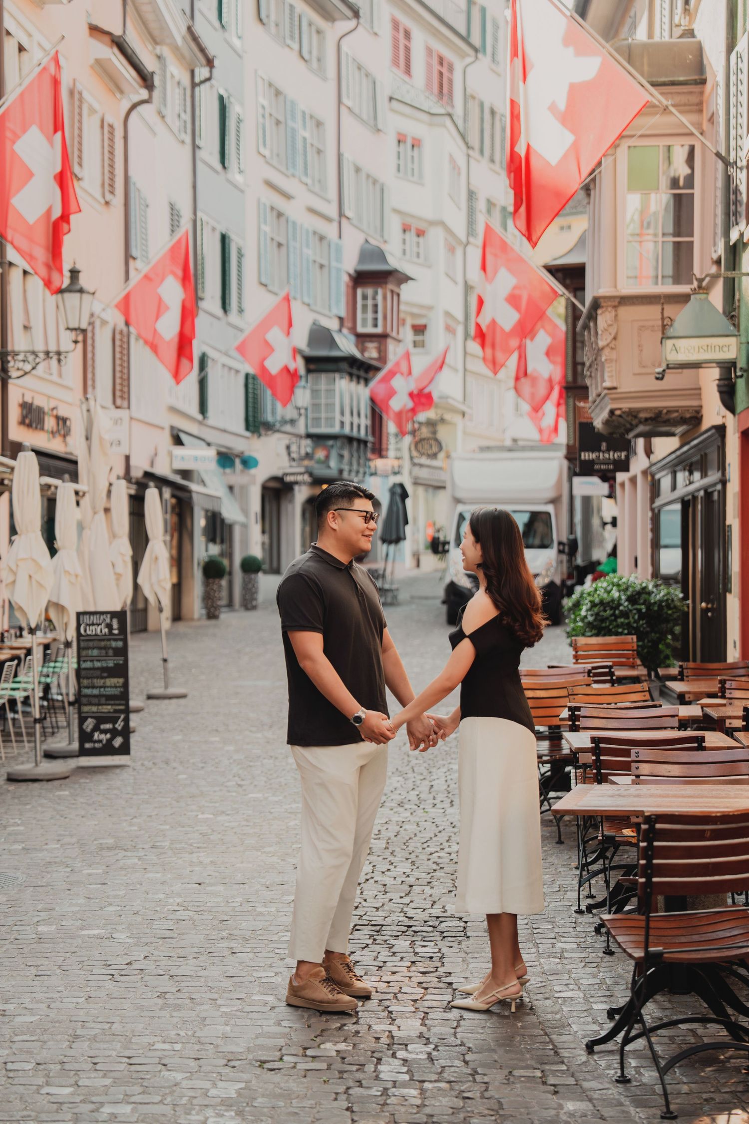 Couple holding hands facing each other on a cobblestone street in Zurich, Switzerland, with Swiss flags overhead.