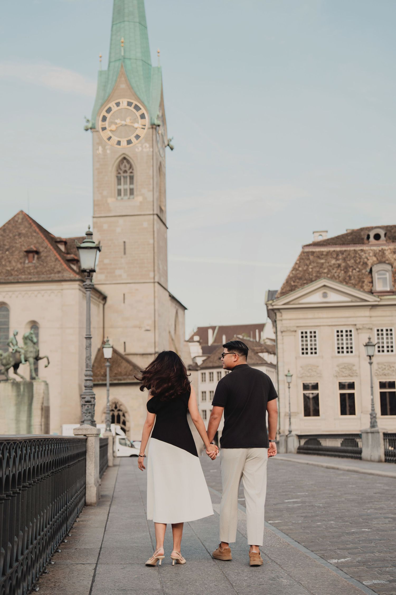 Couple walking hand in hand on a Zurich bridge with Fraumunster clock tower and old town skyline.