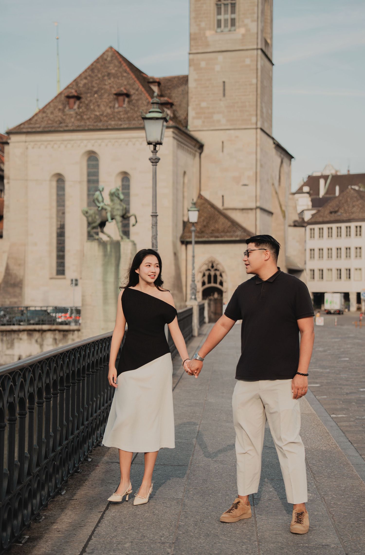 Couple holding hands on a Zurich bridge smiling at each other with historic church in background.