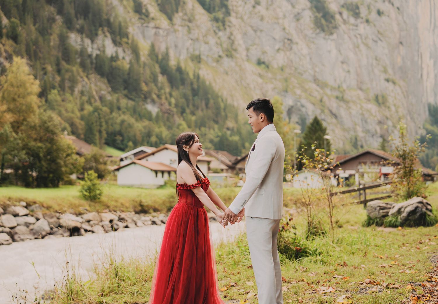 Couple in red bridal gown and grey suit hold hands by rocky riverbank in Swiss mountain valley.