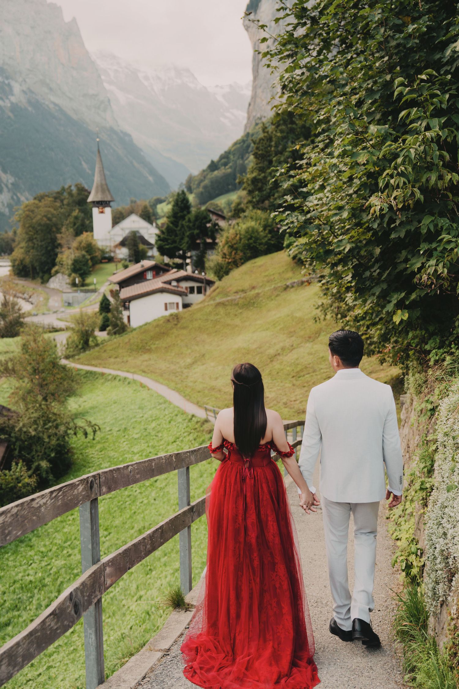 Couple walking hand-in-hand away from camera along scenic Swiss alpine village pathway in formal attire.