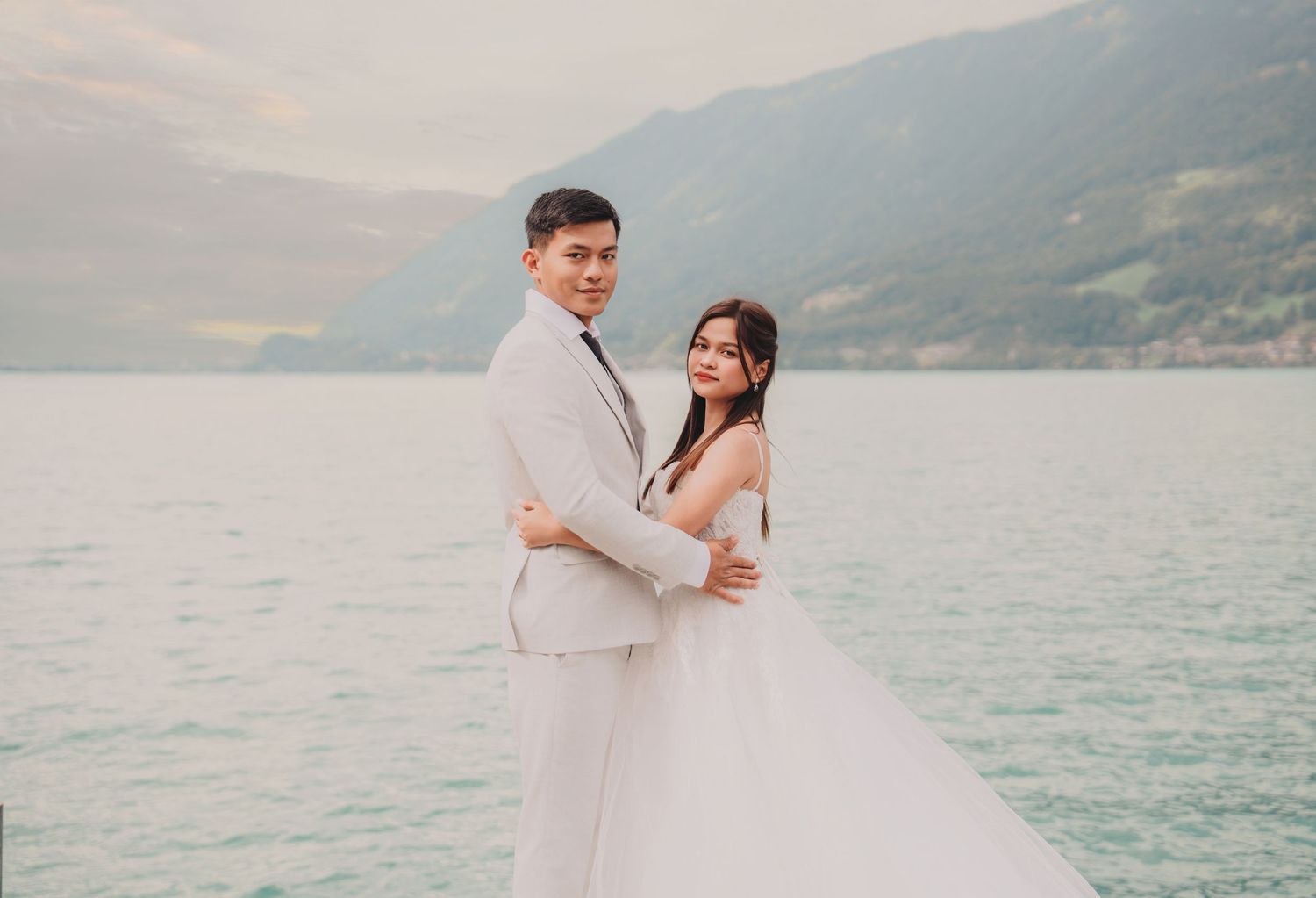 Smiling bride and groom posing together by a serene alpine lake with lush green mountains.