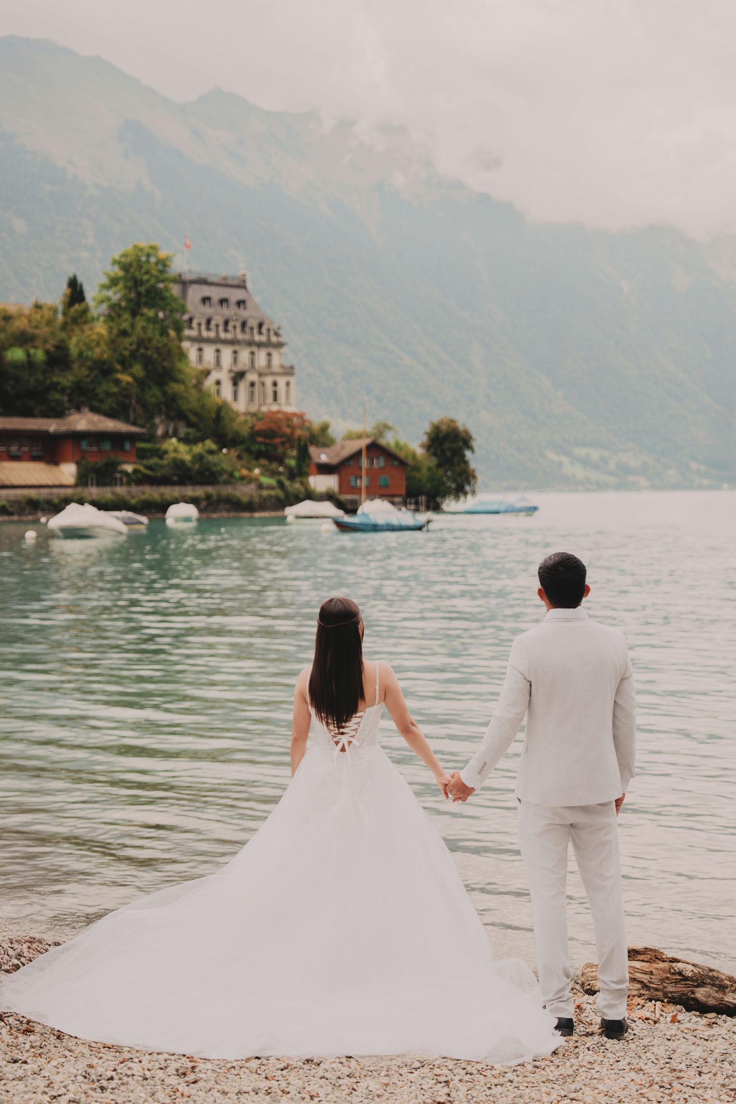 Bride and groom holding hands by a serene alpine lake with mountain views and a historic building in background.