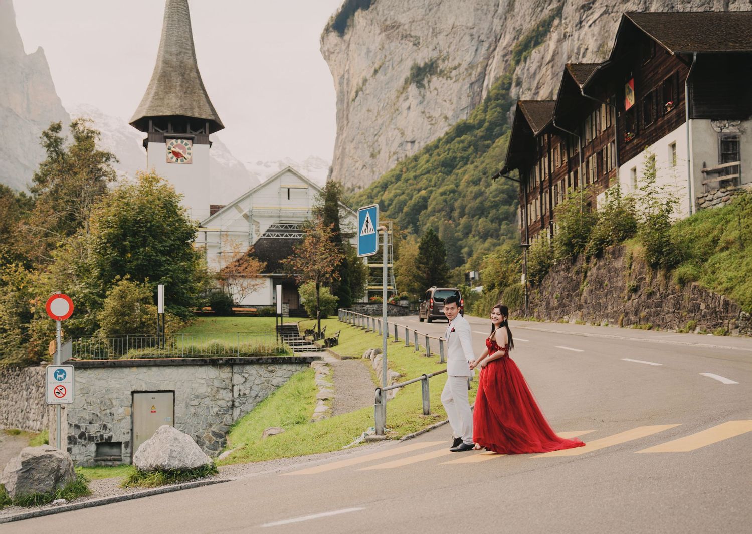 Couple in red dress and white suit crossing road in scenic Swiss alpine village setting.
