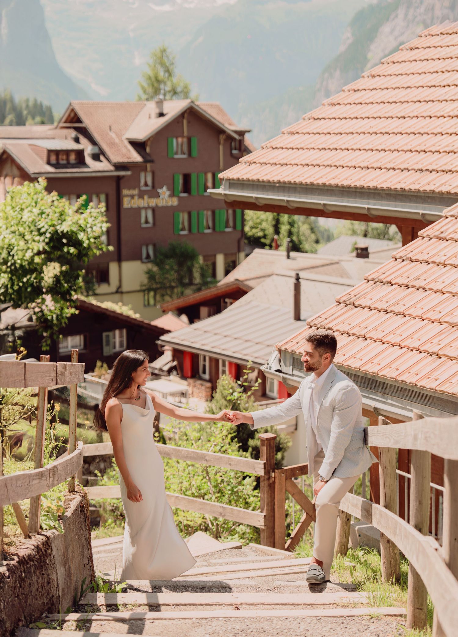 Couple in wedding attire holding hands on wooden stairs in a Swiss Alpine village with mountain backdrop.