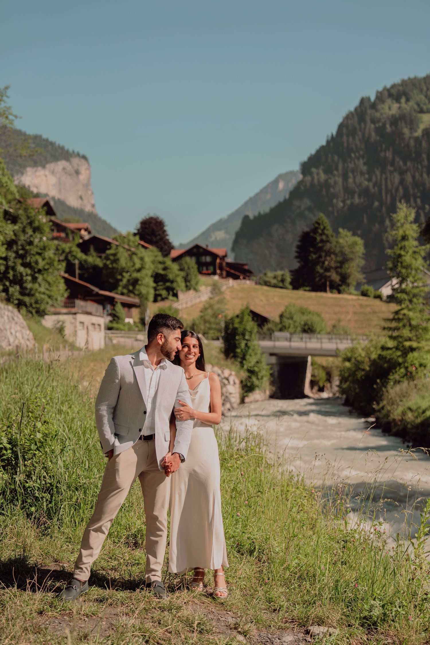 Couple in formal attire standing by a river with Swiss mountain village background.