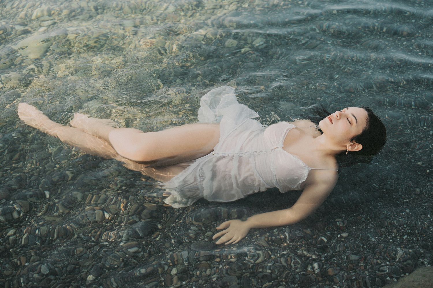 Woman in sheer white dress floating on her back in clear shallow water with eyes closed.