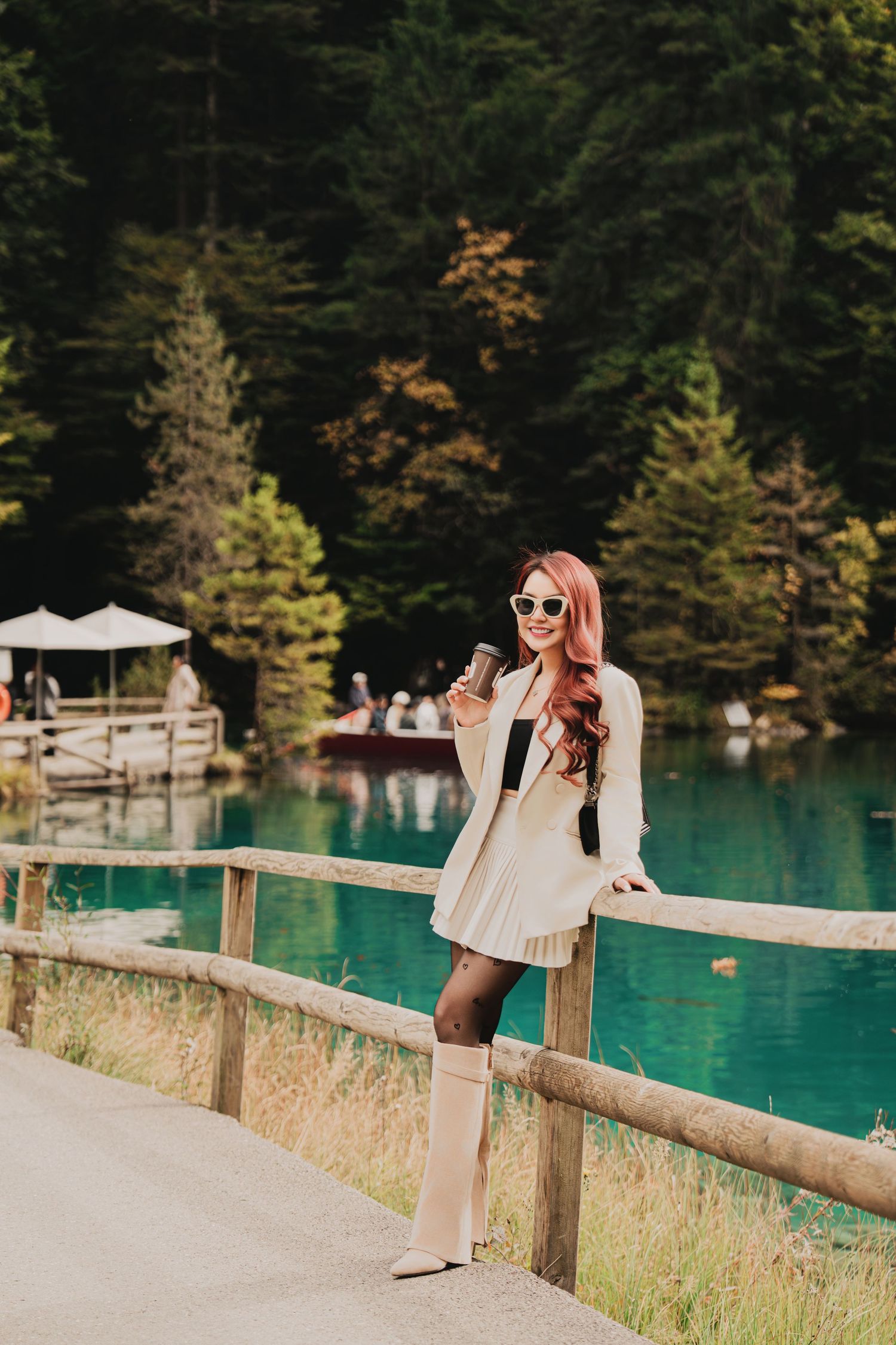 Stylish red-haired woman in white outfit with coffee cup posing by turquoise alpine lake.