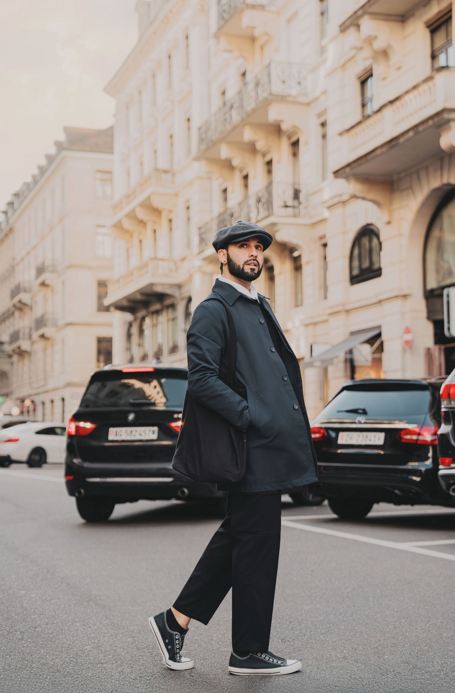 Stylish man in navy coat and newsboy cap walking across urban street with elegant European architecture.