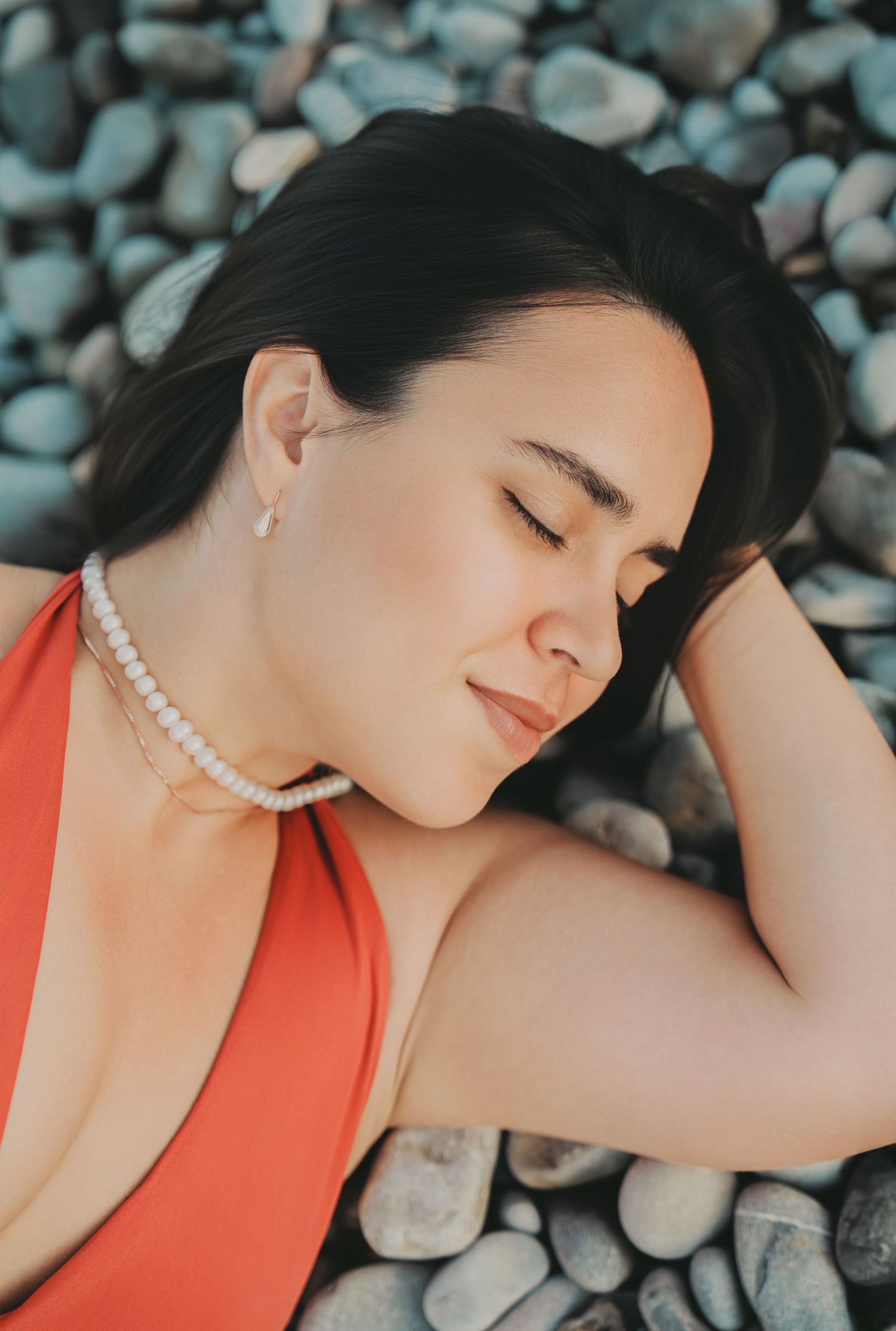 Young woman in red swimsuit wearing pearl necklace resting on rocky beach with eyes closed.