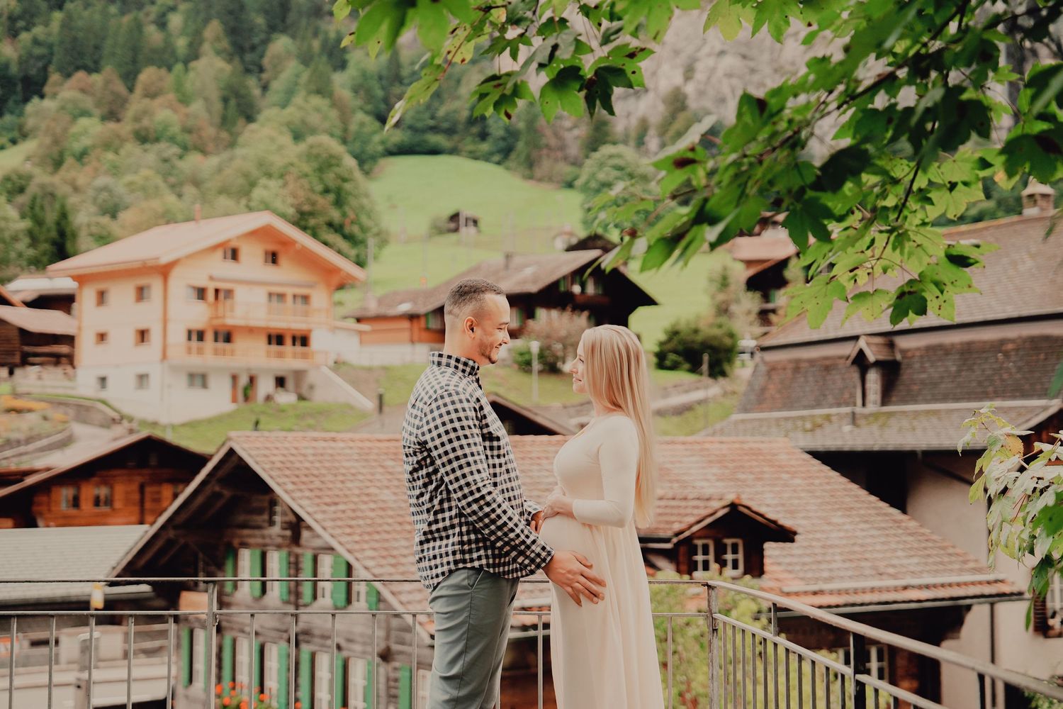 Expectant couple holds hands facing each other on a balcony with Swiss alpine village rooftops below.